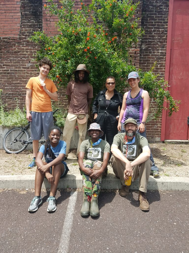 A team of 7 POP and OEEC staff and volunteers at a sunny work day in front one of the site's pomegranate bushes, which grows flush against a brick wall, summer 2021.